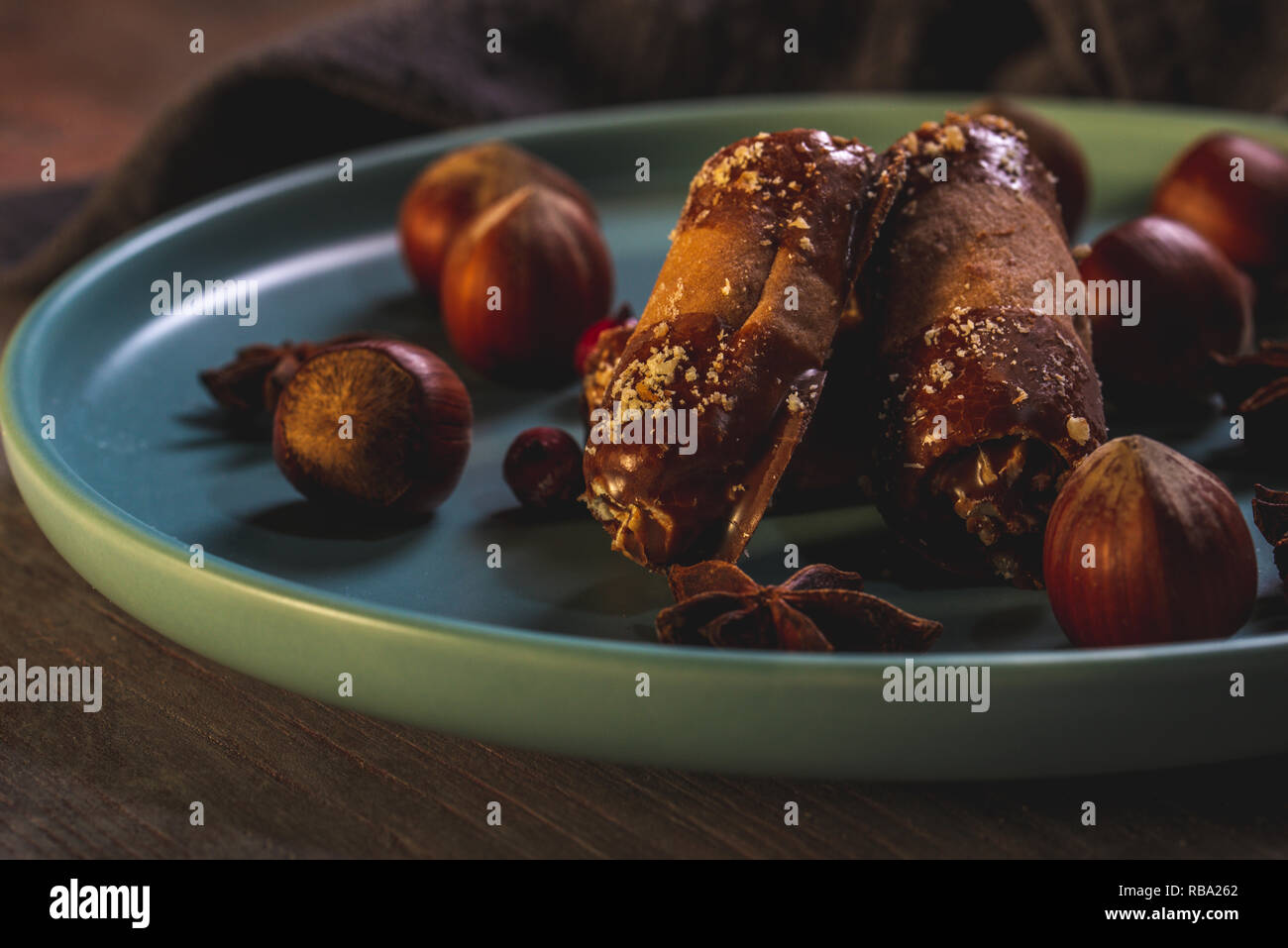 Horizontal photo of few sweet rolls stuffed by cream with chocolate on ends. Sweets are on blue plate with several raw hazelnuts. Plate is on wooden b Stock Photo