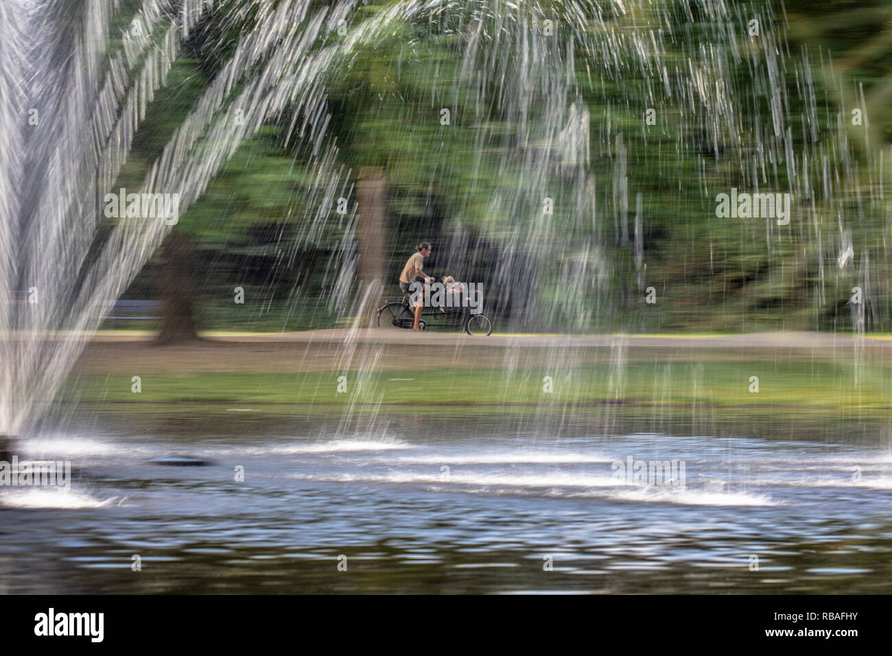 The Netherlands, Amsterdam. Vondelpark. Parent brings his children to school by bicycle or cargo bike. Fountain. Blurred motion. Stock Photo