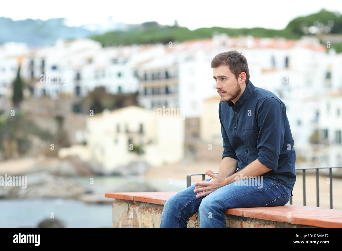 Worried man looking away sitting on a ledge in a coast town Stock Photo