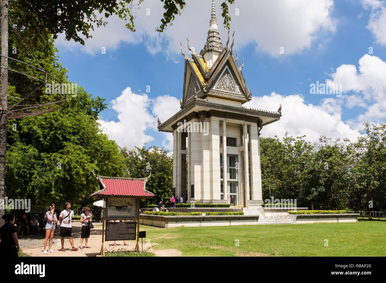 The Killing Fields Genocidal Centre memorial site with Buddhist Stupa containing human skulls and tourists reading sign Choeung Ek Phnom Penh Cambodia Stock Photo