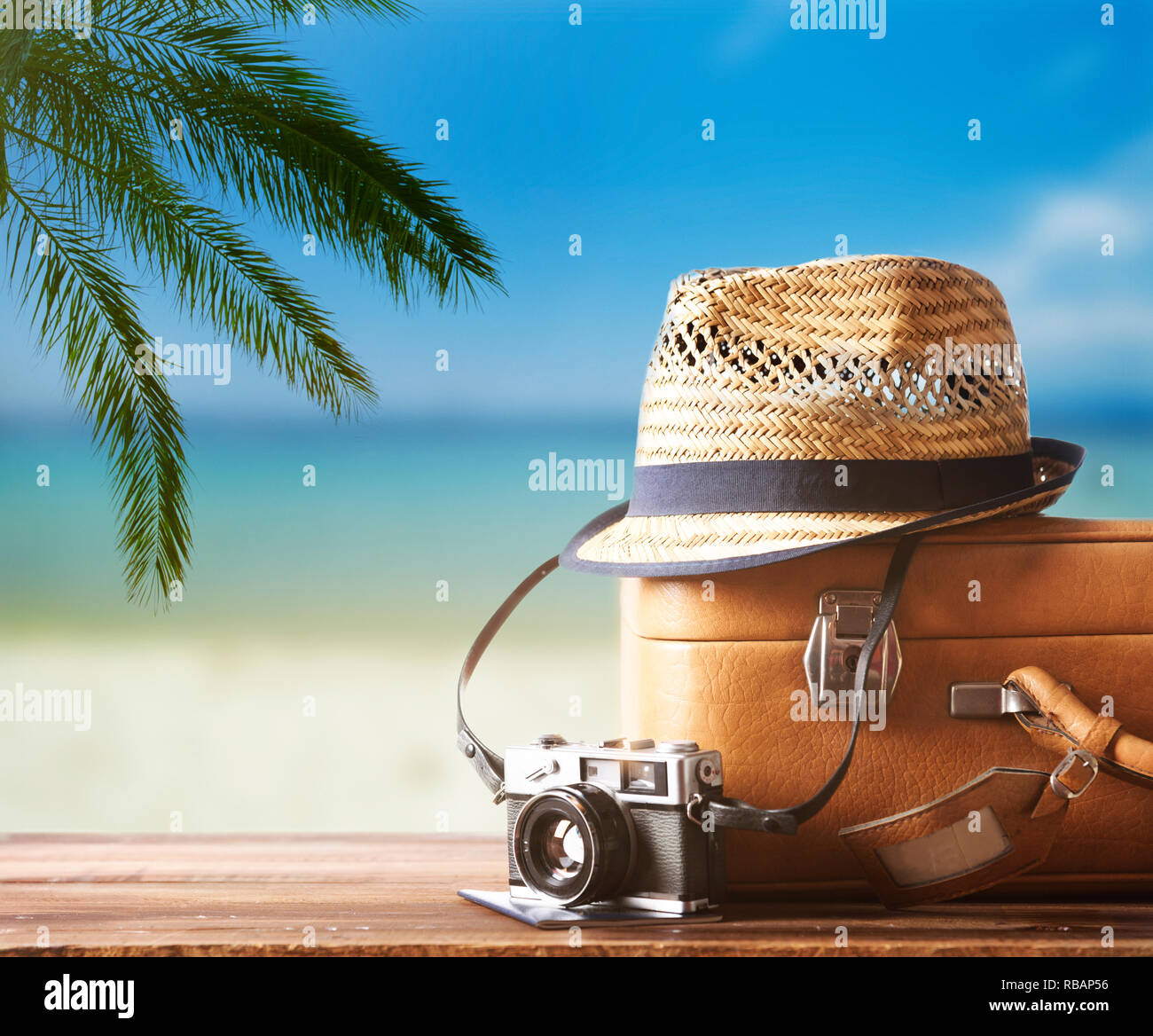 Vintage suitcase, hipster hat, photo camera and passport on wooden dack. Tropical sea, beach and palm three in background. Summer holiday traveling de Stock Photo