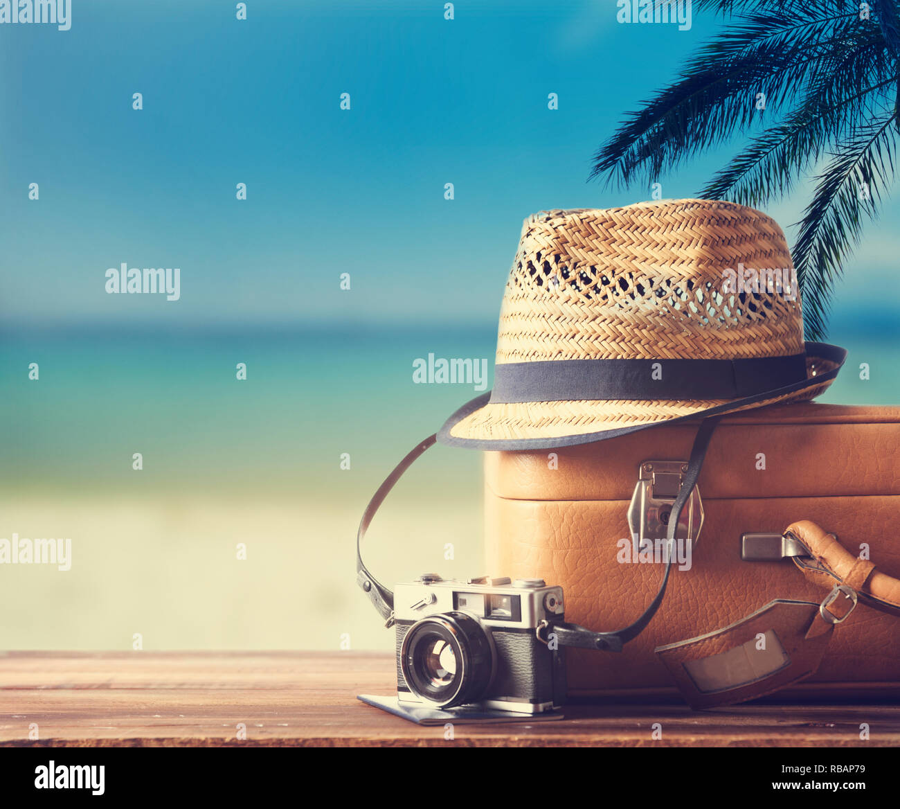 Vintage suitcase, hipster hat, photo camera and passport on wooden dack. Tropical sea, beach and palm three in background. Summer holiday traveling de Stock Photo