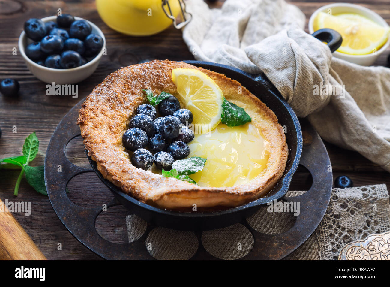Fresh homemade Dutch Baby pancake with lemon curd and blueberries in iron skillet on rustic wooden background. Stock Photo