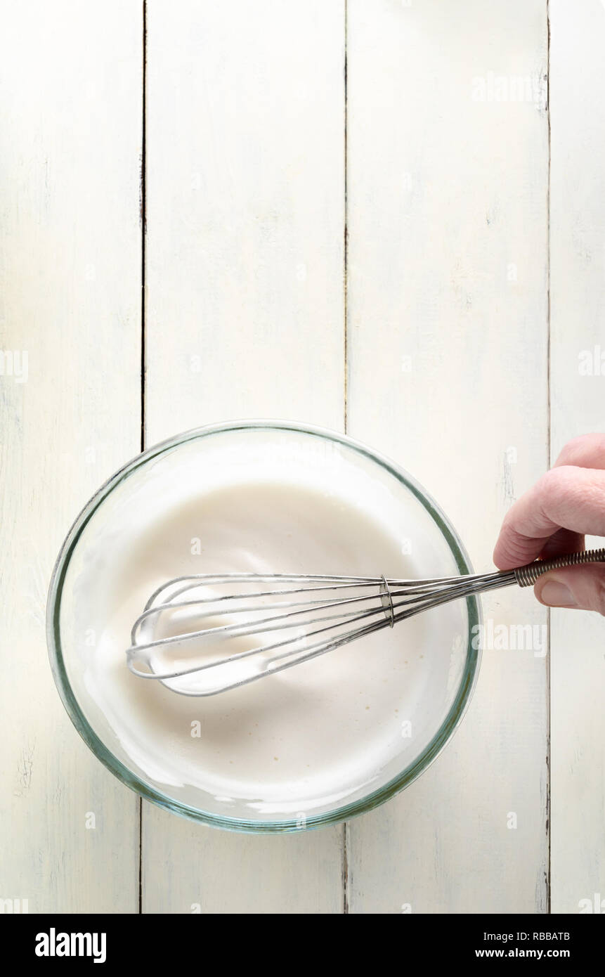 Overhead shot of frothy aquafaba (chickpea water). A vegan egg white replacer in glass bowl with hand holding metal whisk on painted white wood planke Stock Photo