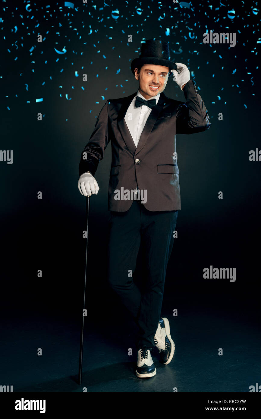 Professional showman wearing suit gloves and top hat standing isolated on black background holding cane posing to camera looking aside smiling playful Stock Photo