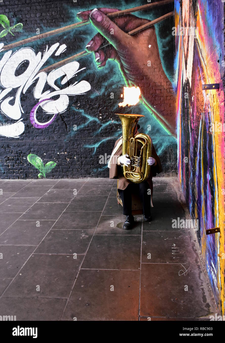 A street musician plays a tuba with fire close to the Borough Market in front of a wall with graffiti. London, South Bank, United Kingdom. Stock Photo