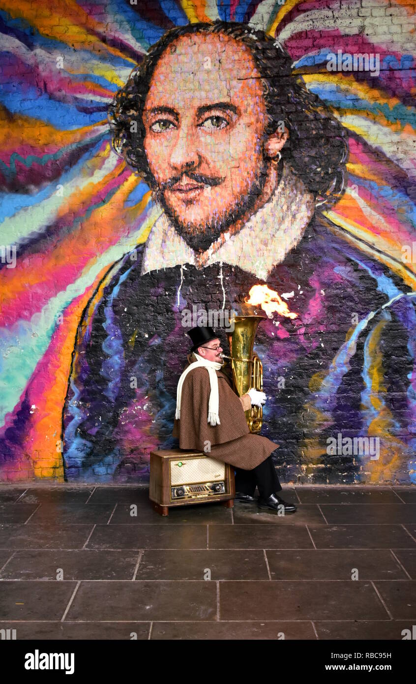 A street musician plays a tuba with fire close to the Borough Market in front of a wall with graffiti. London, South Bank, United Kingdom. Stock Photo