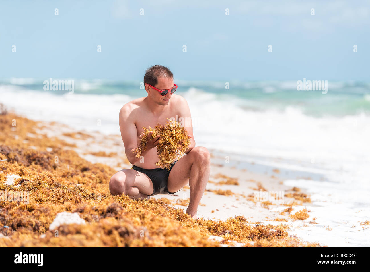 Young man on beach during sunny day with red sunglasses in Miami, Florida with blue sky background, sitting squatting crouching holding yellow sargass Stock Photo