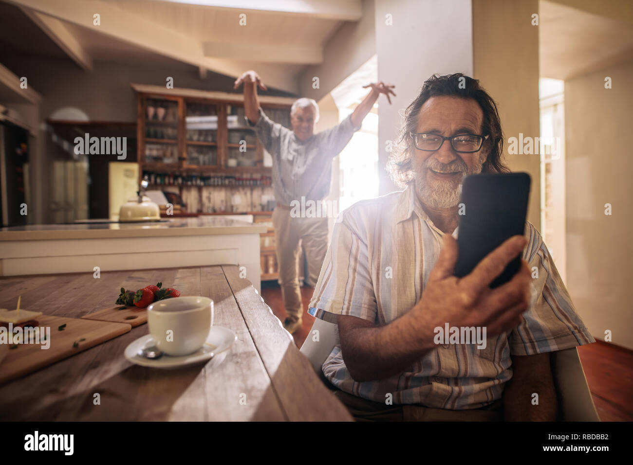 Happy senior man taking selfie with friend standing at back making funny gestures.  Two elderly men taking selfie with mobile phone at home. Stock Photo
