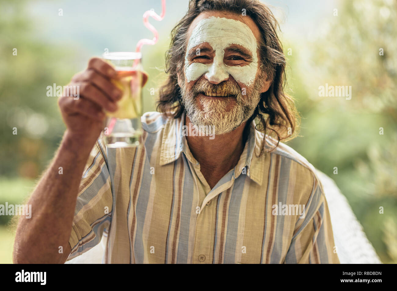 Bearded senior man with clay facial mask holding a glass of juice in hand. Man having spa facial treatment raising his juice glass at camera. Stock Photo