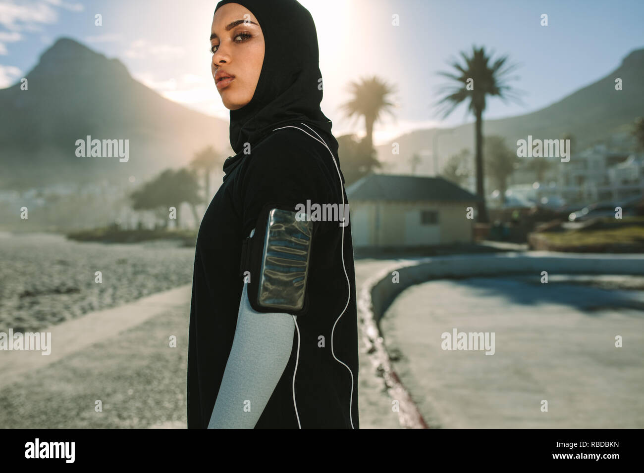 Islamic woman in sportswear standing outdoors in city on sunny day. Muslim female runner in hijab listening to music from mobile phone and looking at Stock Photo