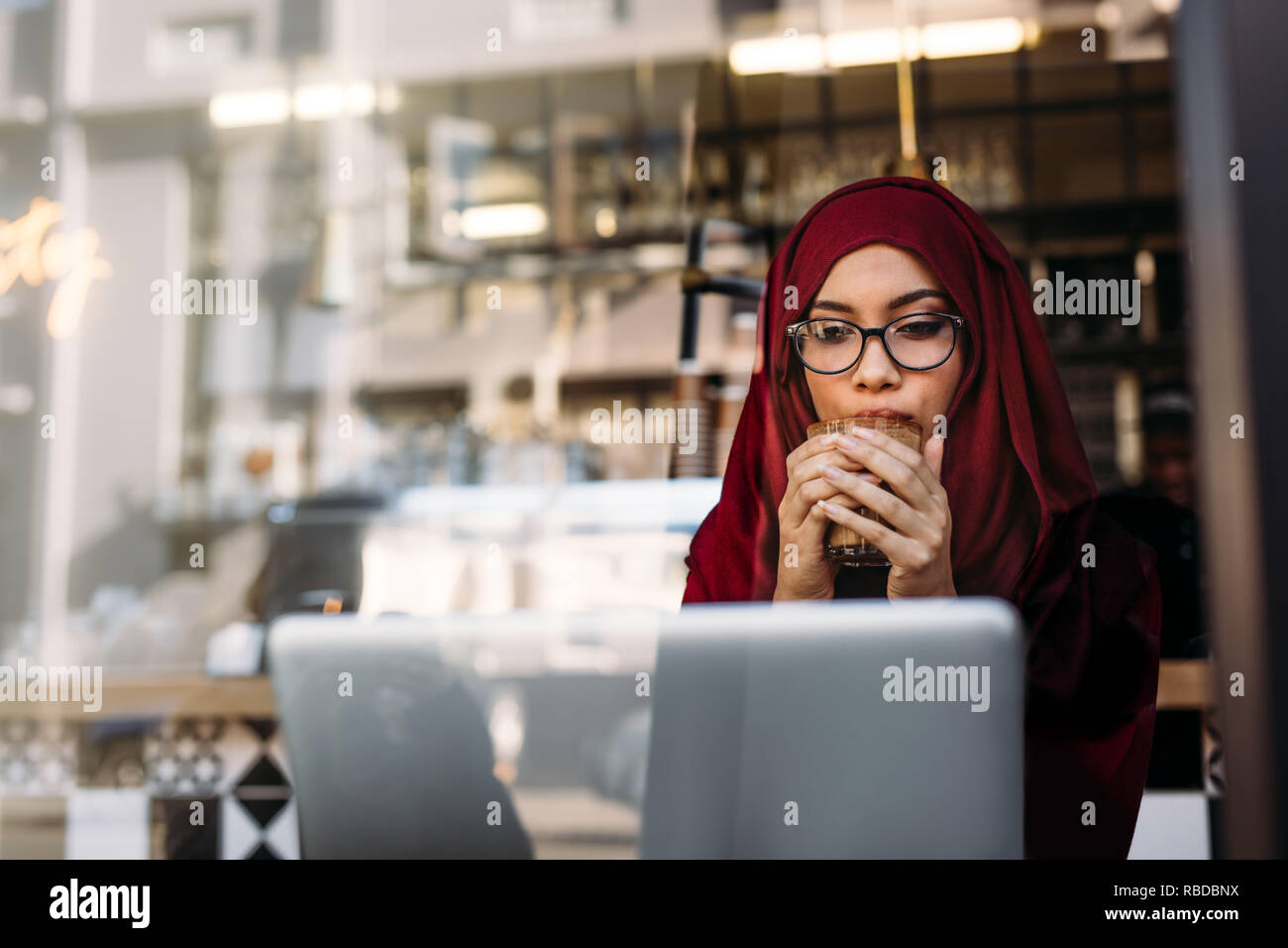 Muslim woman with hijab looking laptop and drinking coffee sitting at cafe. Islamic female having morning coffee at cafe and using laptop computer. Stock Photo