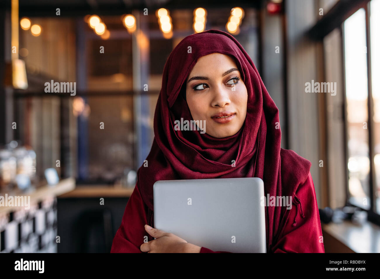 businesswoman in hijab with laptop at cafe looking away. Young muslim woman in hijab standing at restaurant. Stock Photo
