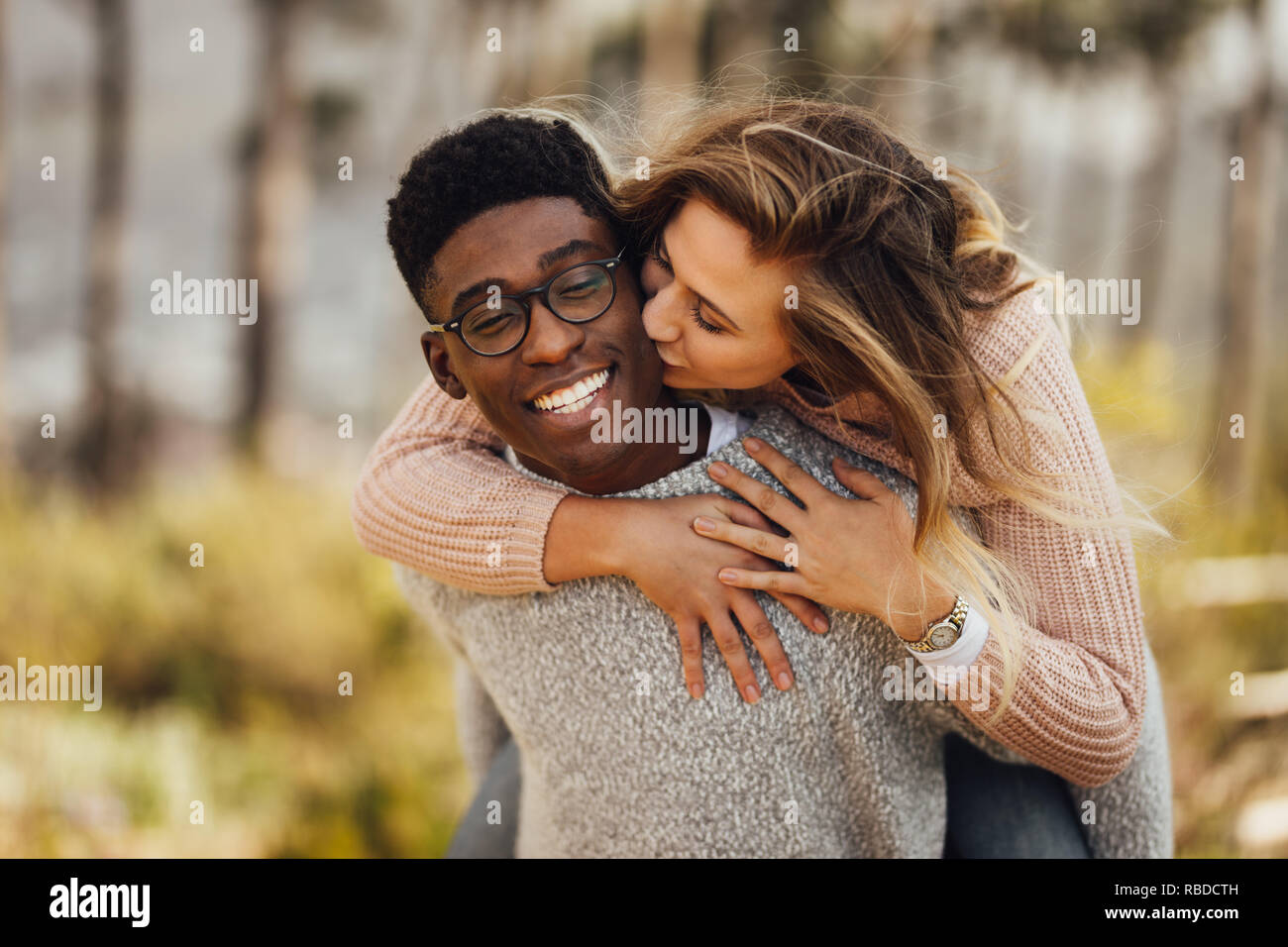 Handsome young man giving piggyback ride to his girlfriend. Couple having fun outdoors. Man carrying his girlfriend on his back, with woman kissing ma Stock Photo