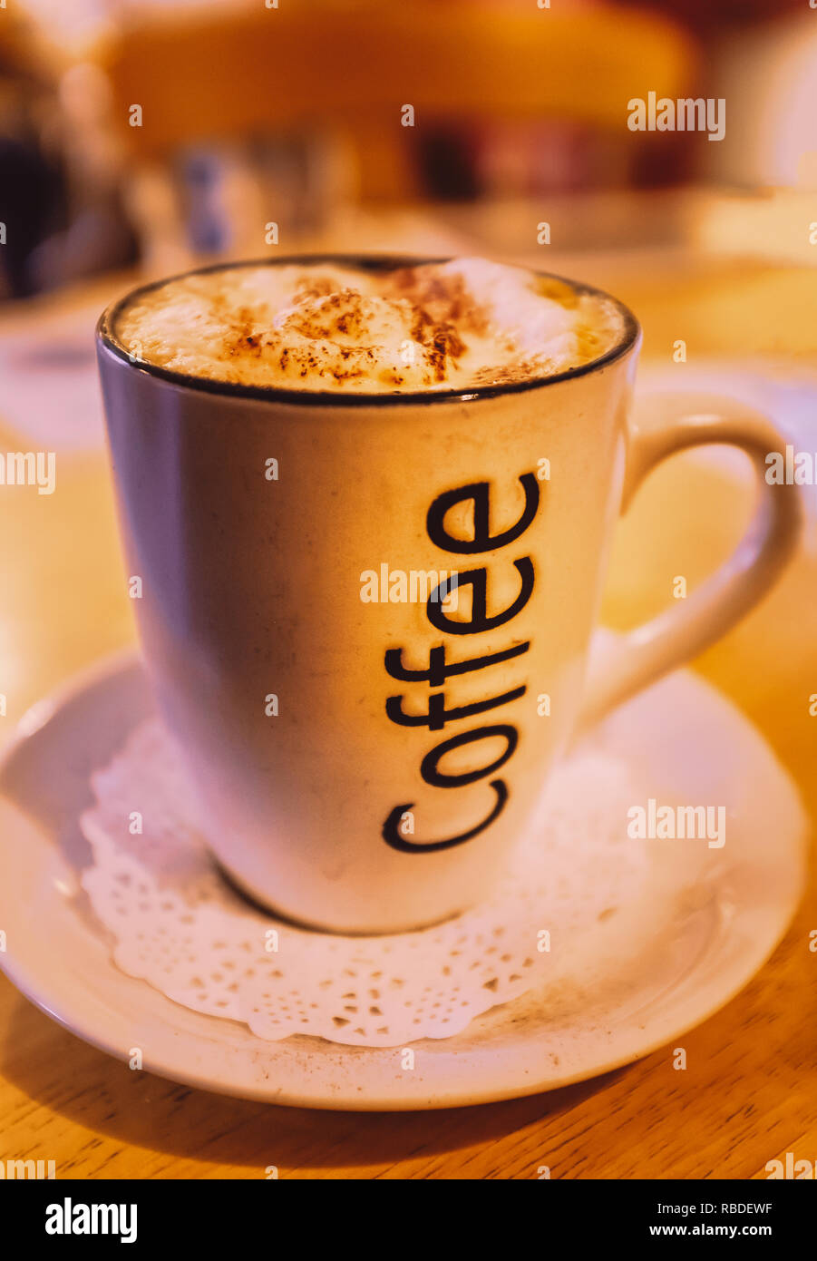 Mug of cappuccino coffee with the word coffee written on the side. On a saucer with a paper lace doily on a round wooden table with a wood chair with  Stock Photo