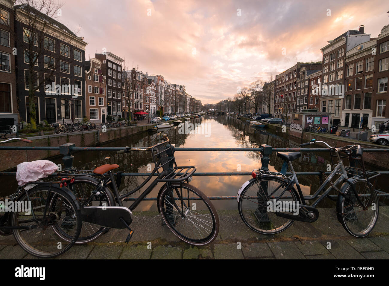Bicycles locked to the bar of a bridge over a canal lined by traditional architecture houses at sunset in the center of Amsterdam, the Netherlands. Stock Photo