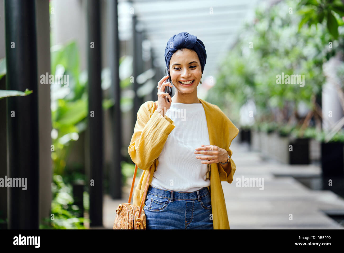 A young and beautiful Middle Eastern woman in a turban hijab is smiling as she talks on her smartphone on the street during the day. Stock Photo