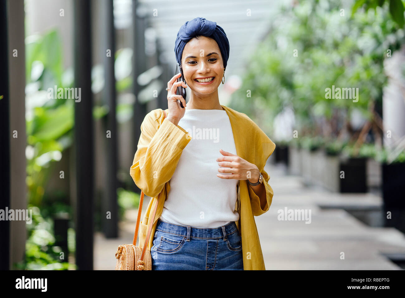 A young and beautiful Middle Eastern woman in a turban hijab is smiling as she talks on her smartphone on the street during the day. Stock Photo