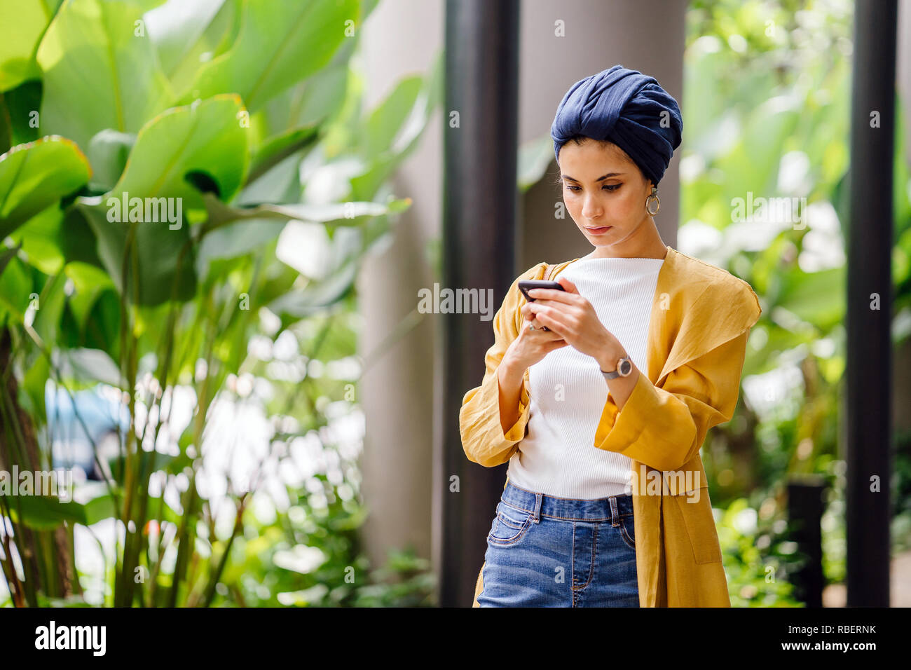 A Middle Eastern Arab woman in a headscarf is texting and messaging on her smartphone on a street in the city during the day. Stock Photo