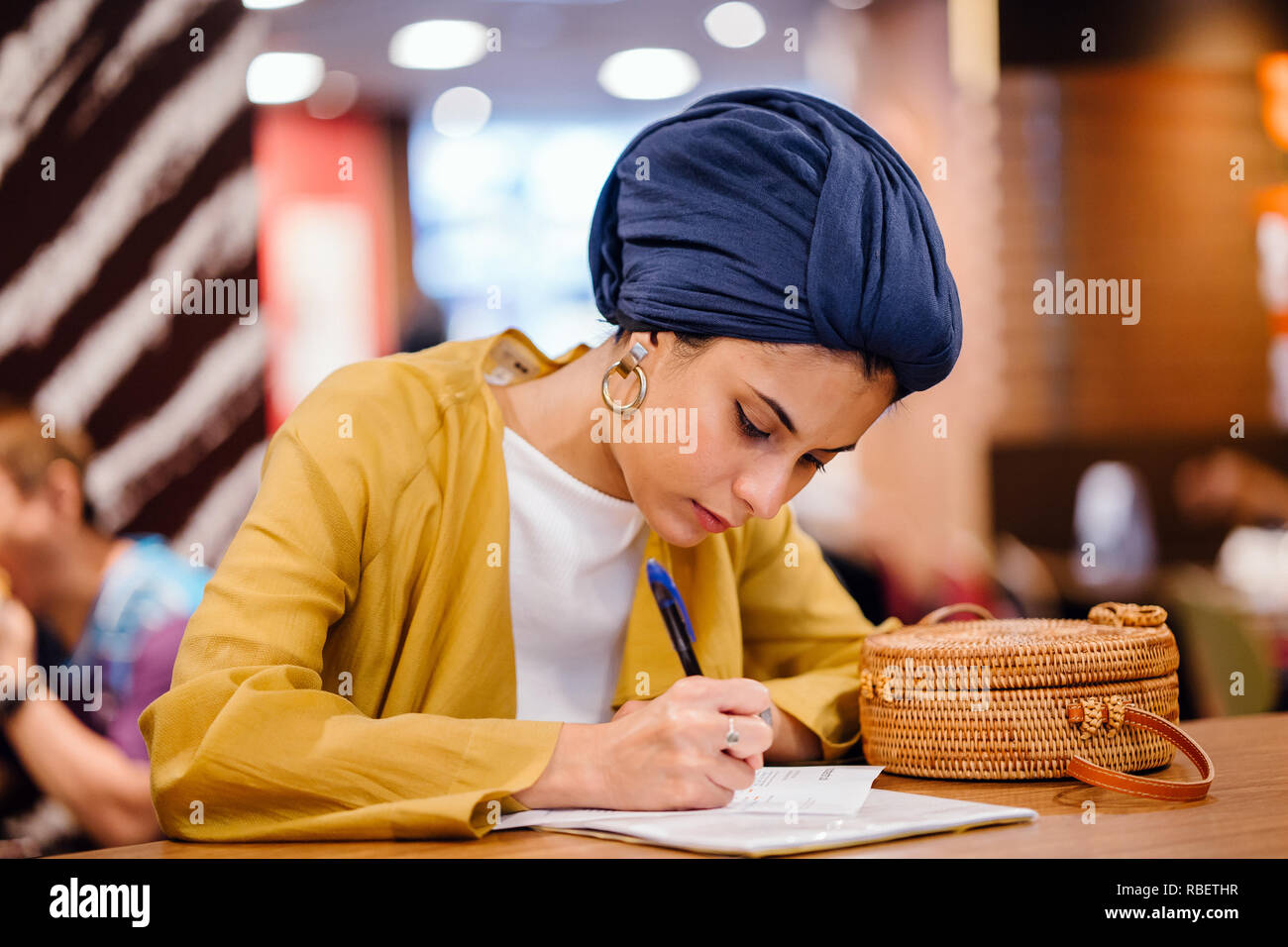 Portrait of a beautiful and elegant Middle Eastern woman writing in her journal notebook. She is wearing a blue turban hijab headscarf. Stock Photo