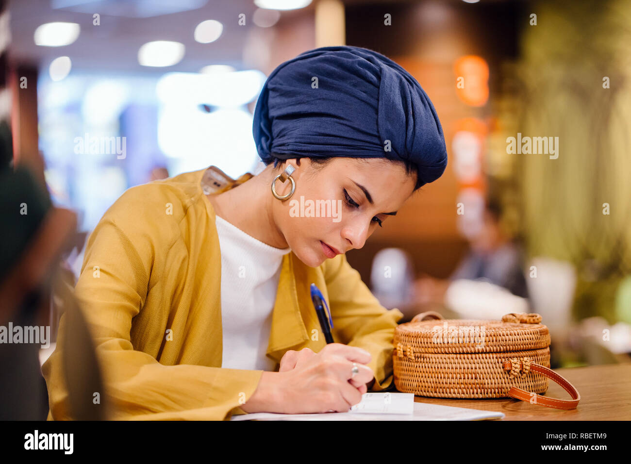 Portrait of a beautiful and elegant Middle Eastern woman writing in her journal notebook. She is wearing a blue turban hijab headscarf. Stock Photo