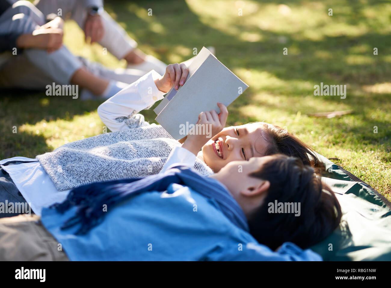 two little asian kids boy and girl lying on back on grass in park reading a book together with parents sitting in background. Stock Photo