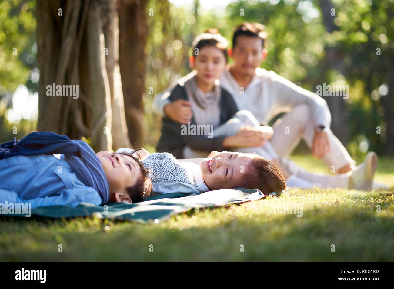 two asian children little boy and girl having fun lying on grass with parents sitting watching in background. Stock Photo