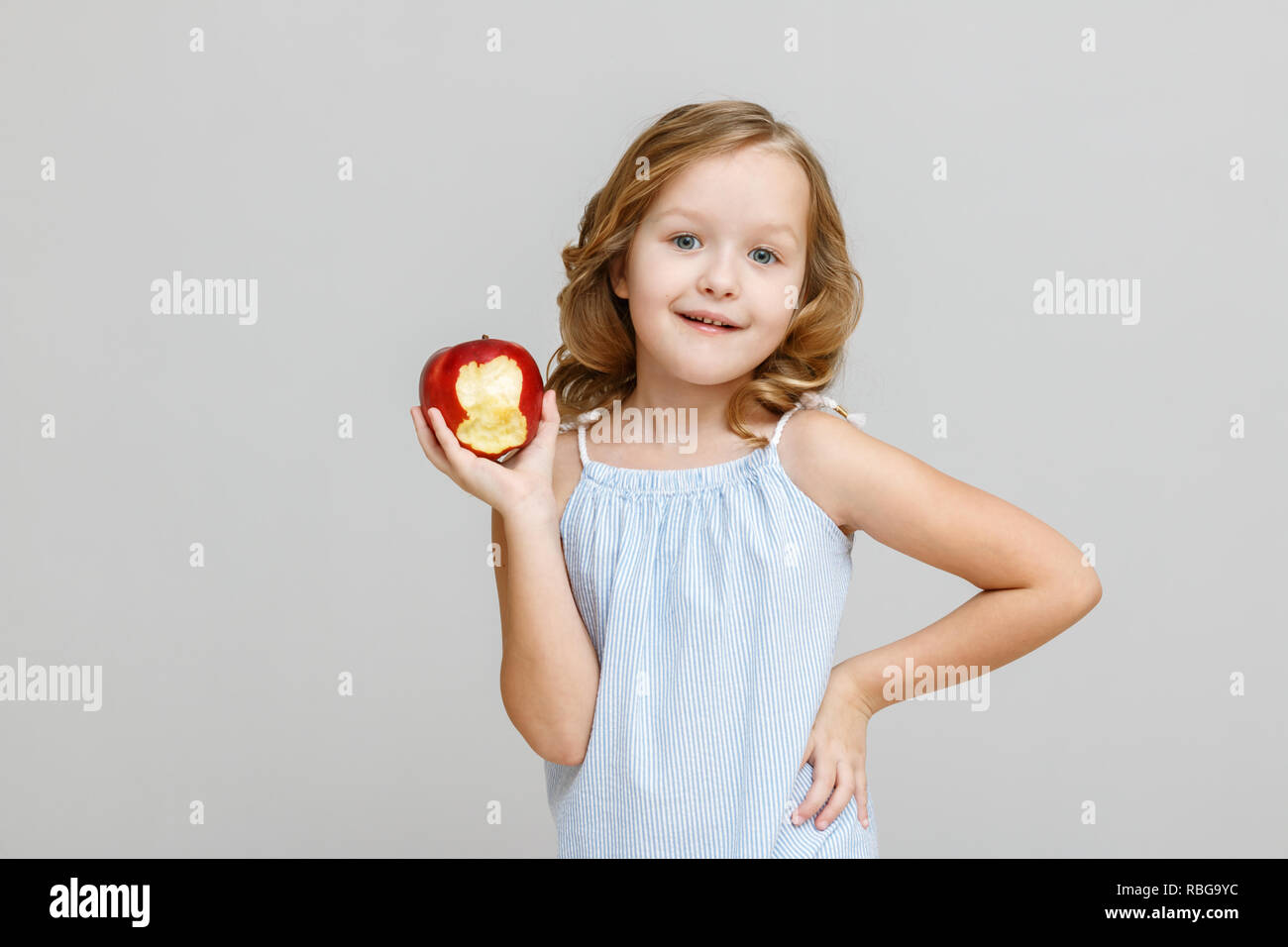 Portrait of a happy smiling little blonde child girl with a bitten red apple on a gray background Stock Photo