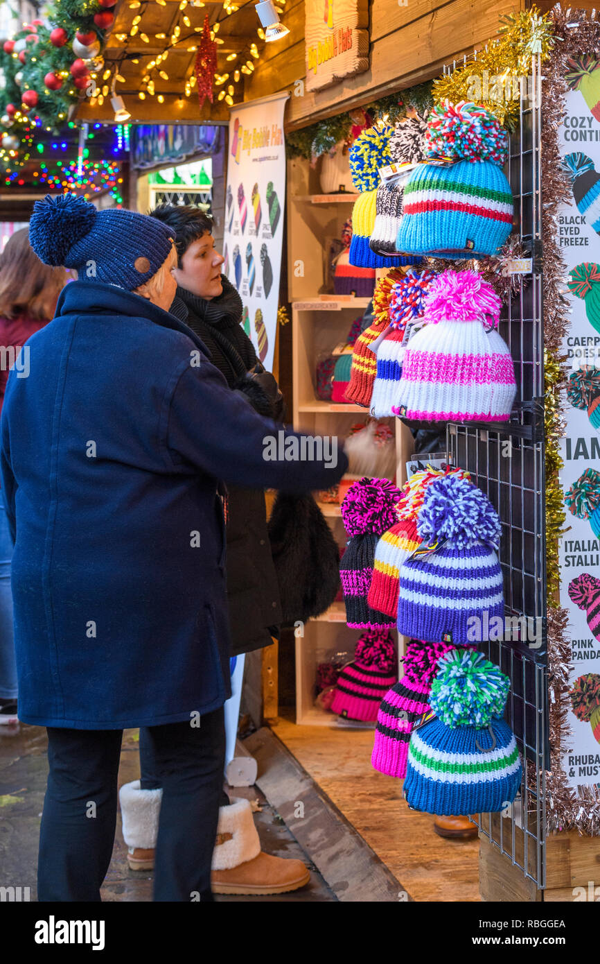 People (women) at Christmas Market stall (potential customers) looking at variety of colourful woolly bobble hats on display - York, England, GB, UK Stock Photo