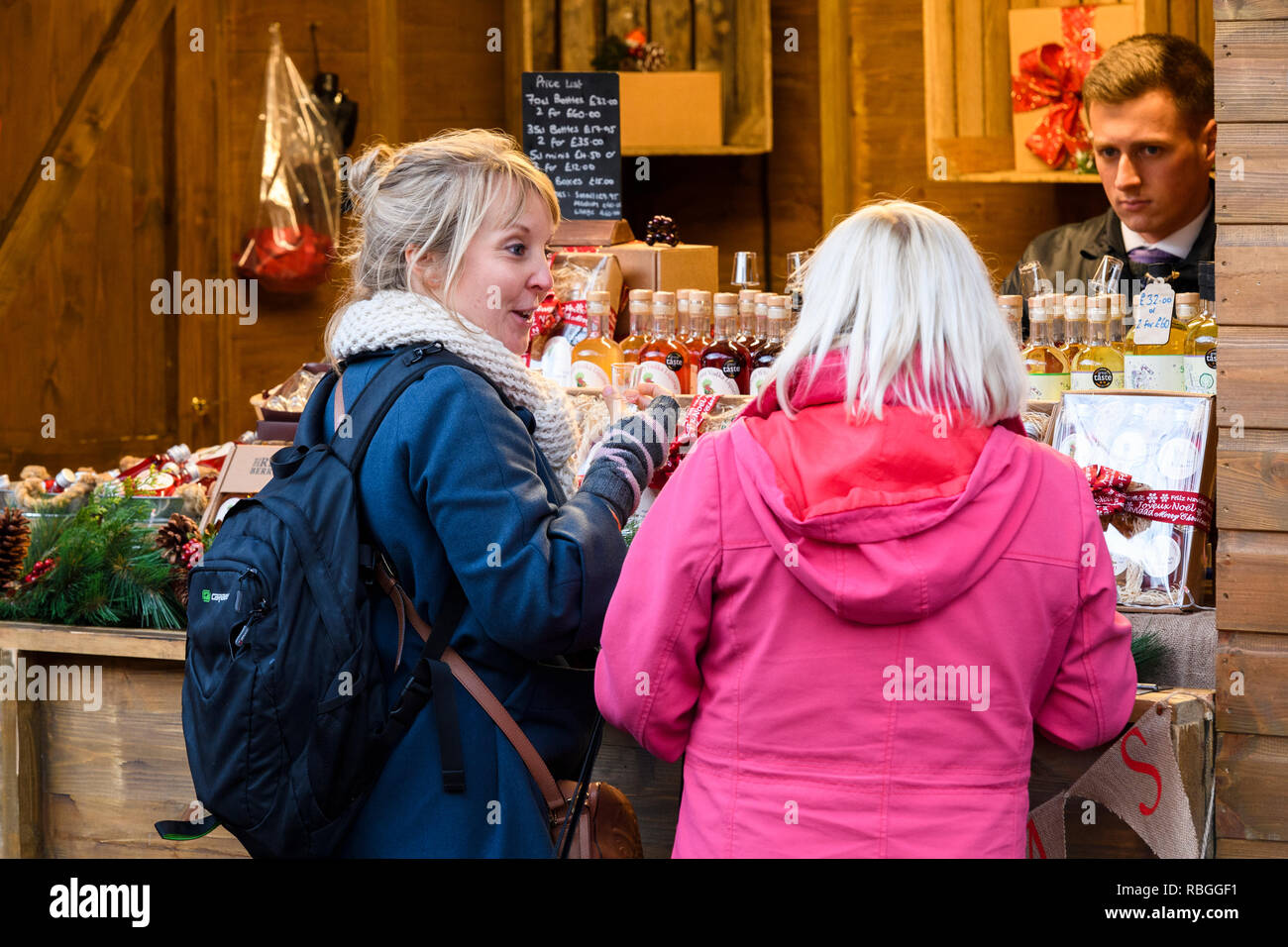 Man working at stall selling alcoholic fruit liqueurs & potential customers (women pals) enjoying sample drinks - York Christmas Market, England, UK. Stock Photo