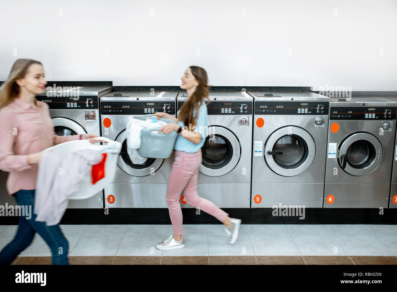Two women walking with clothes at the self service laundry with washing machines on the background, motion blurred effect Stock Photo