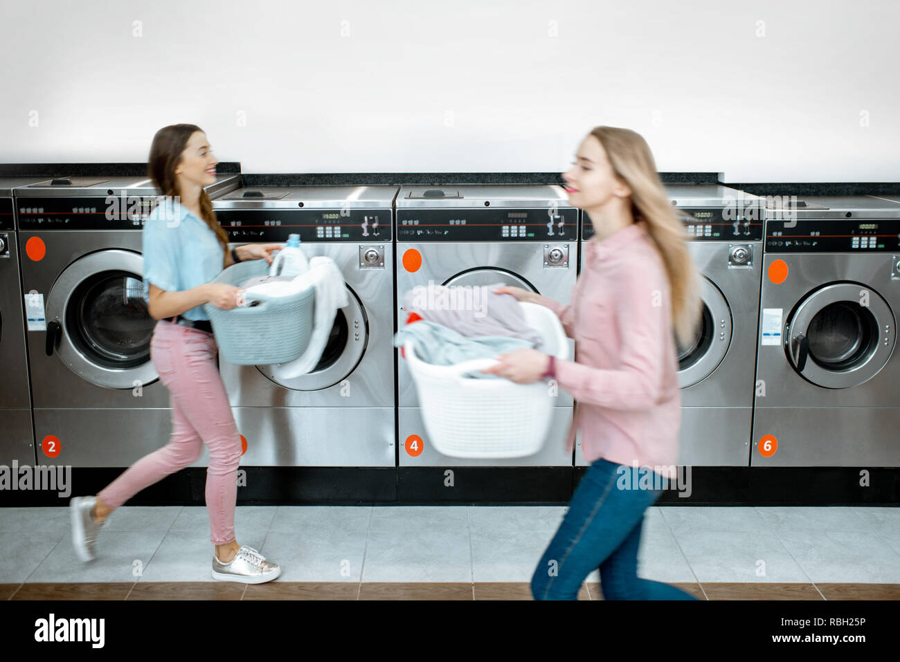 Two women walking with clothes at the self service laundry with washing machines on the background, motion blurred effect Stock Photo