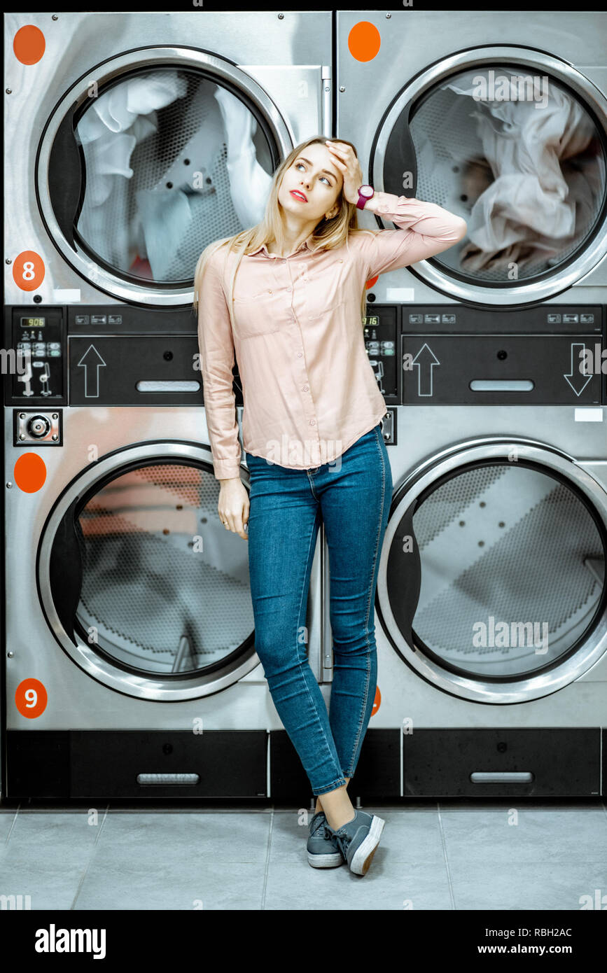 Young woman with boring emotions standing near the dryer machines waiting for clothes to be dried in the laundry Stock Photo