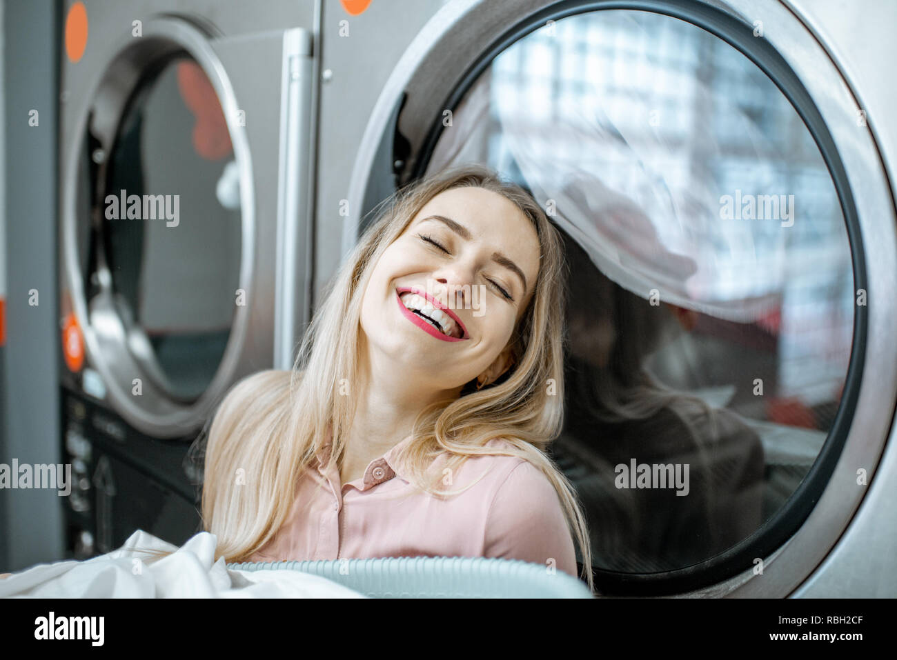 Young and cheerful woman enjoying the washing process standing near professional drying machine in the laundry Stock Photo