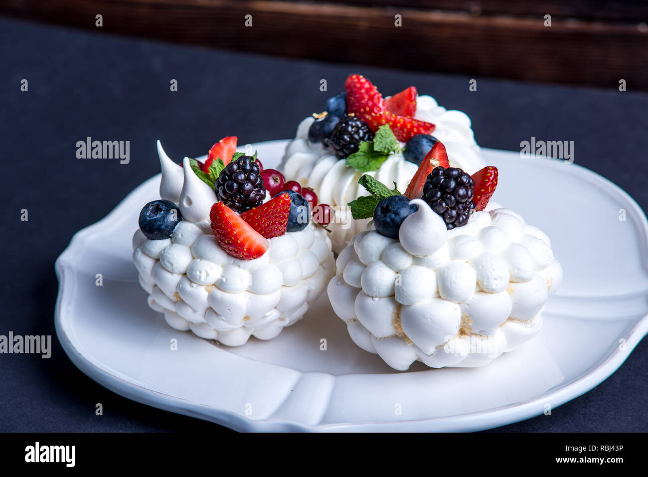 Delicate white meringues with fresh berries on the plate. Dessert Pavlova close-up. Dark background. A festive wedding cake. Stock Photo