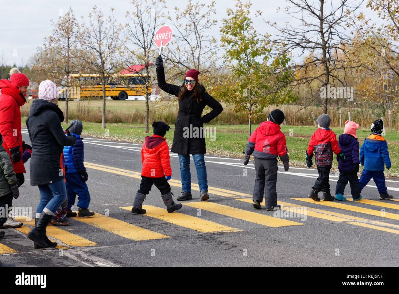 A laughing kindergarten teacher holding up a 'Stop' sign while her pupils cross the road. Photo taken in Quebec so the Stop sign reads Arret. Stock Photo