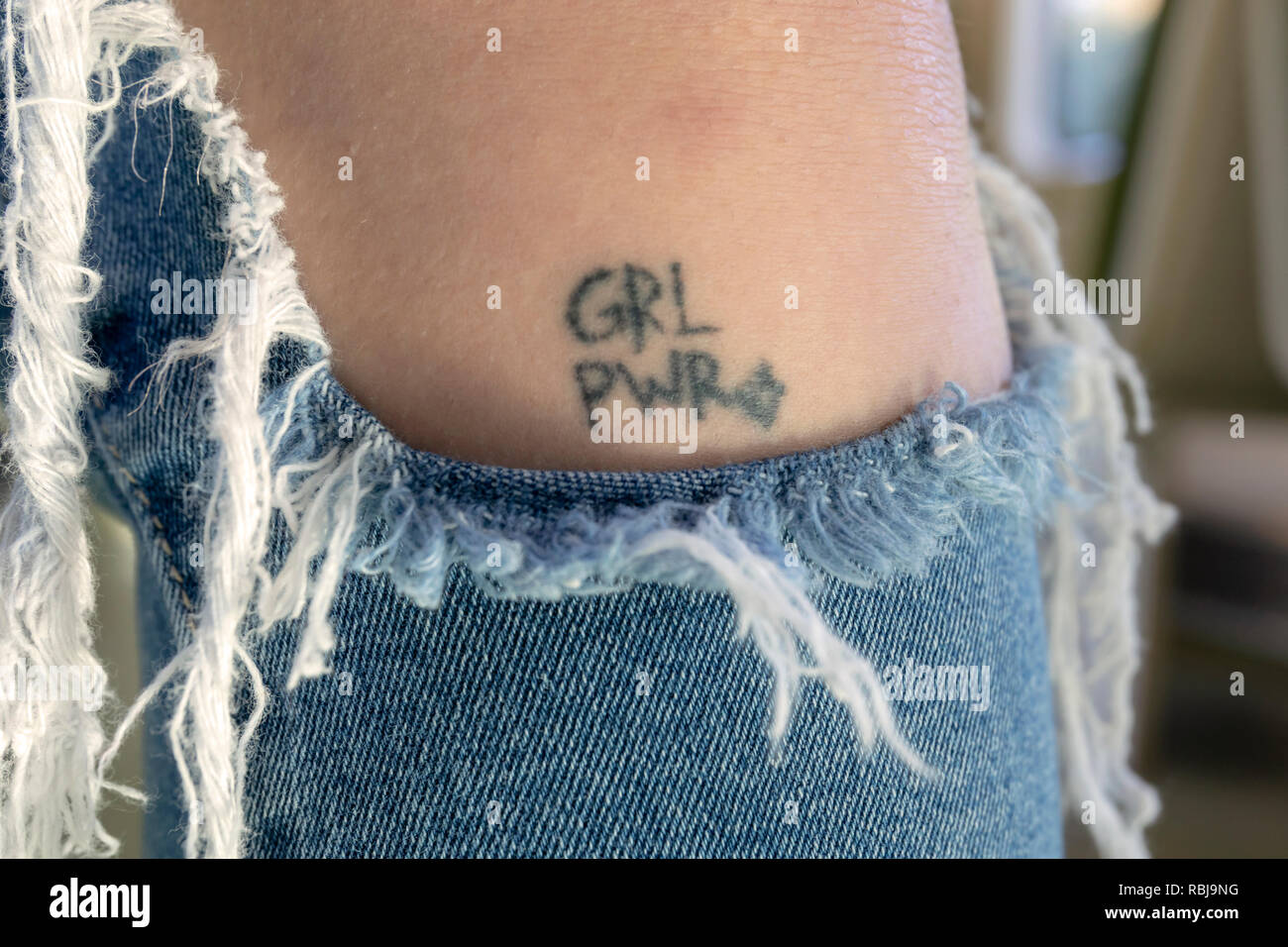 A young woman wearing torn jeans displaying a 'Girl Power' tattoo on her leg. Stock Photo