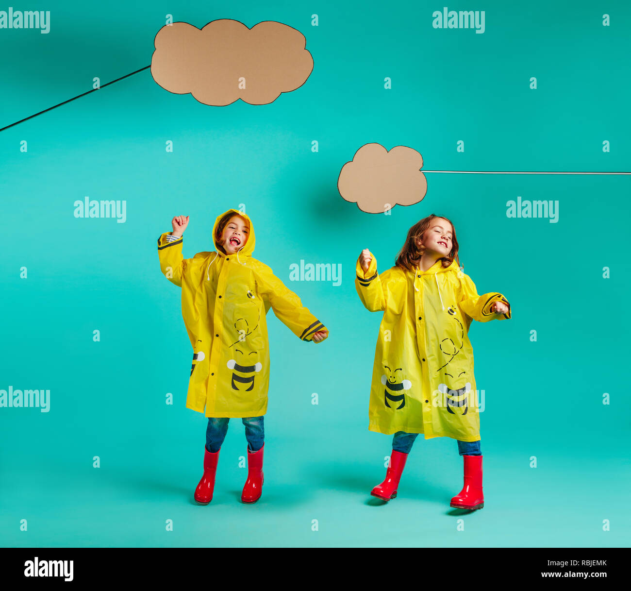 Cute twins girls dancing in raincoats under paper clouds. Girls in rainwear dancing on blue background. Stock Photo