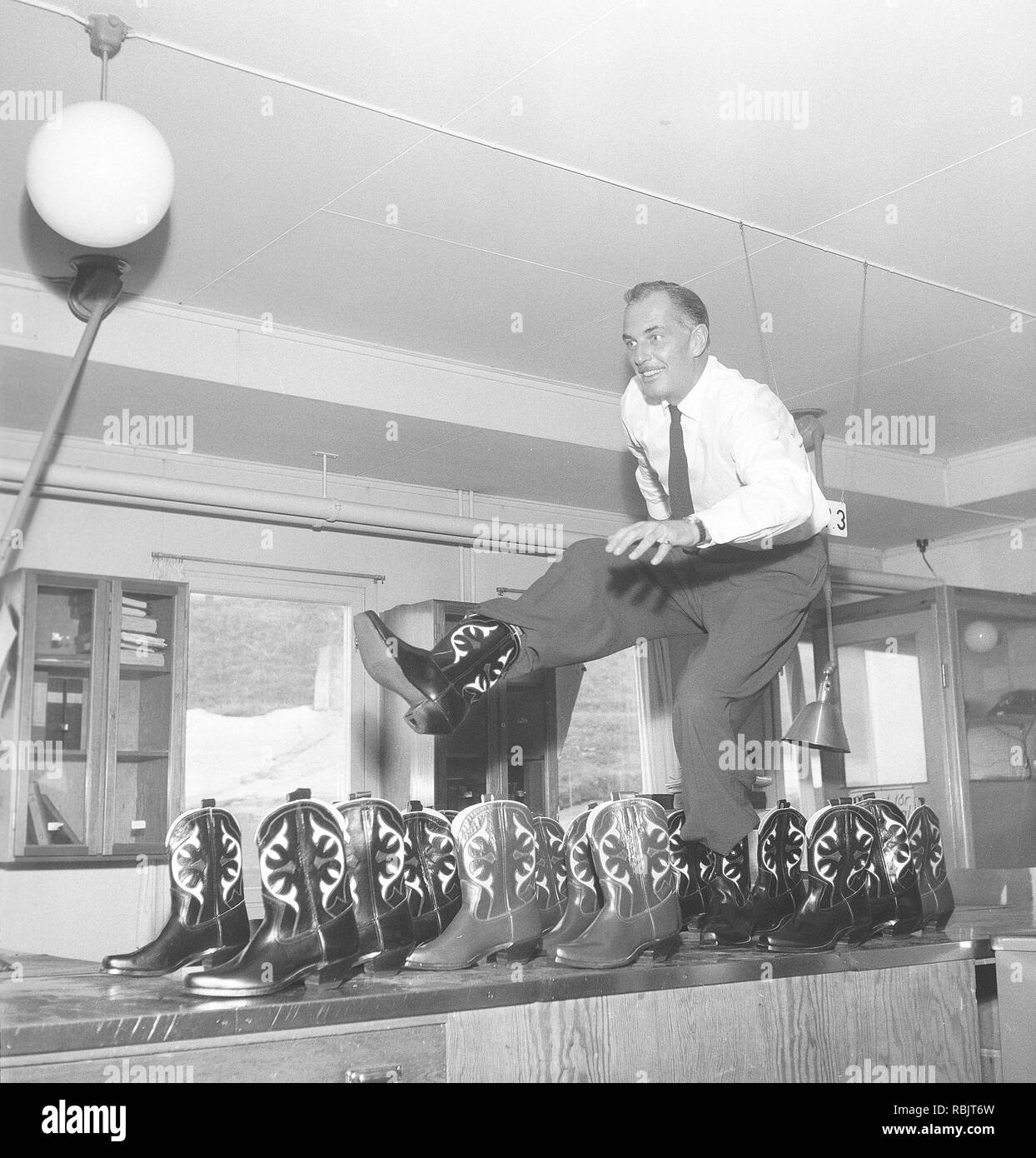 Cowboy boots. A man is playing around and dancing on a table with cowboy boots. Another dozen of cowboy boots are standing on the side. Sweden 1950s. Photo Kristoffersson Ref BX20-6 Stock Photo