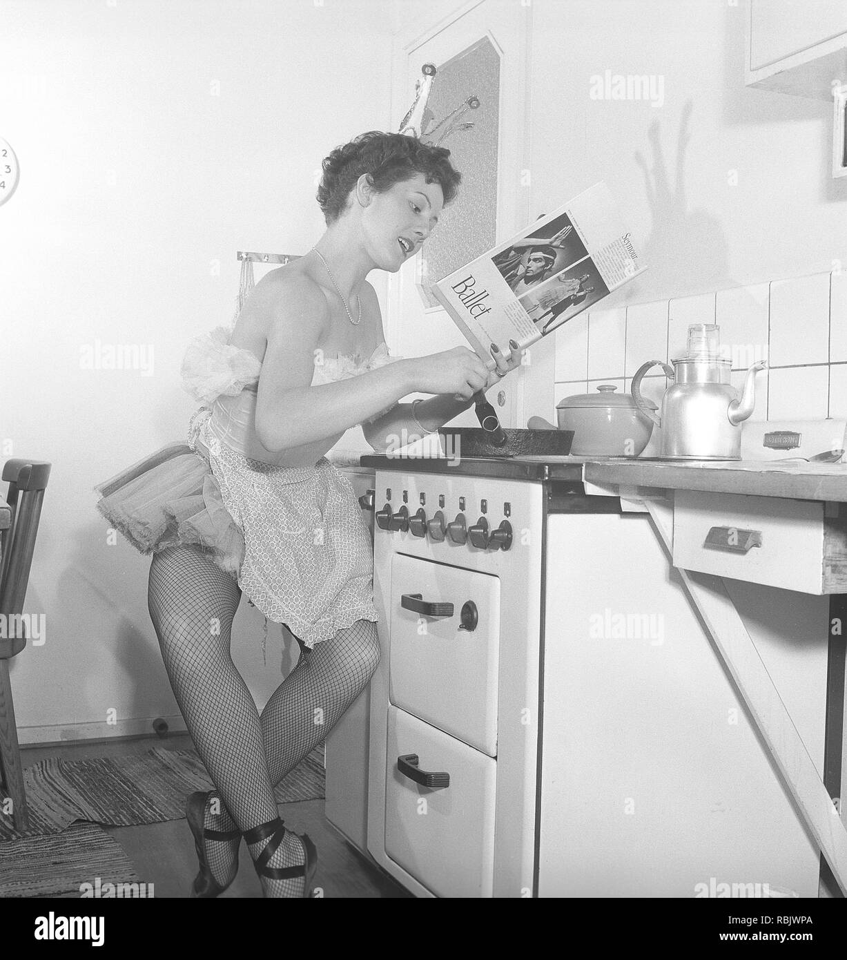 Ballerina in the 1950s. A young ballerina all dressed up in her costume and shoes is sitting by the kitchen stove reading a book about Ballet. Photo Kristoffersson Ref BK5-4. Sweden 1953 Stock Photo