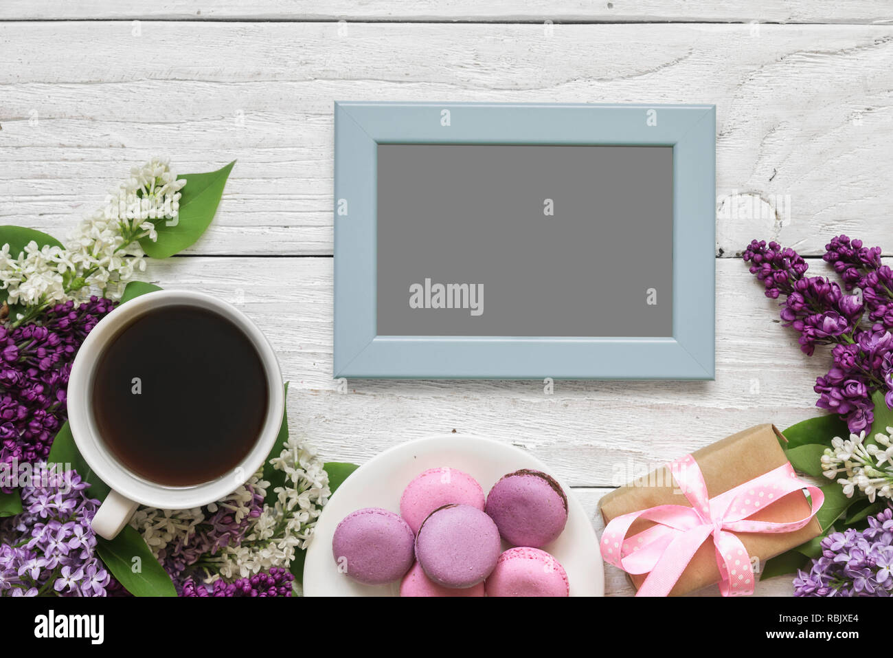 blank photo frame with lilac flowers, coffee cup, sweet macarons and gift box on white wooden background. top view with copy space. mock up Stock Photo