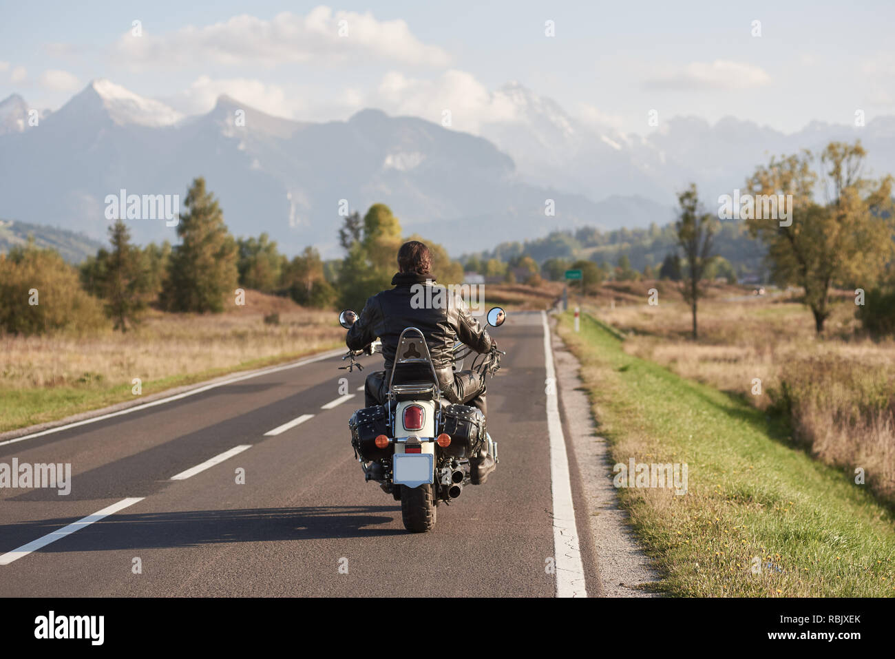 Back view of biker in black leather jacket riding motorcycle along road on blurred background of beautiful mountain range with snowy peaks, moving veh Stock Photo