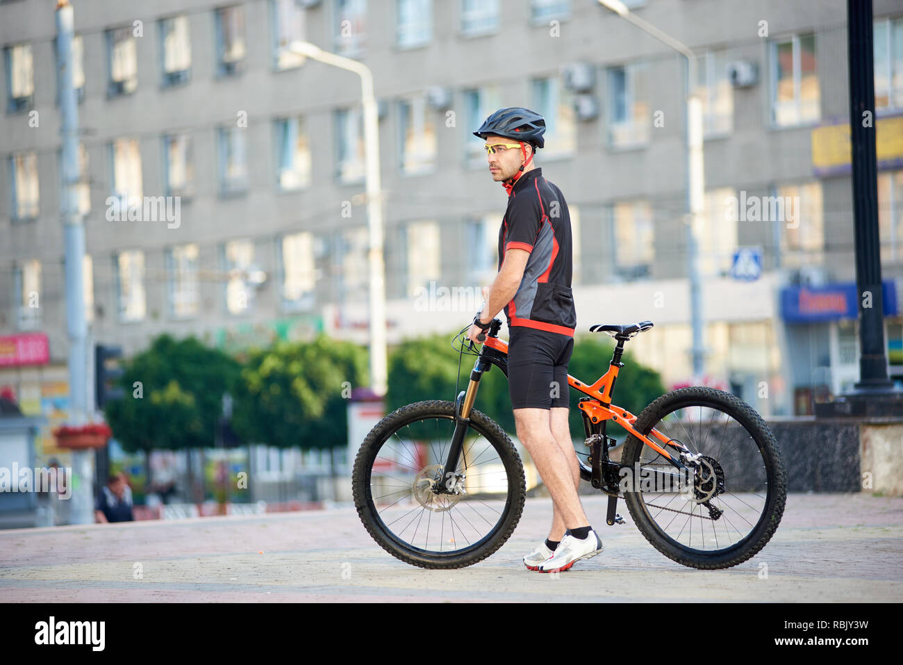 Young man in relaxing after bike ride standing near bicycle in front of city building. Cyclist looking far and posing for sport garment ad campaign. Concept of healthy lifestyle Stock Photo