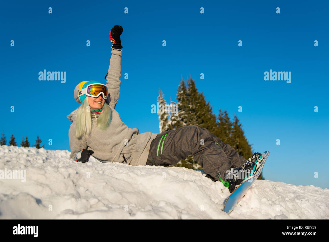 Low angle shot of a happy girl snowboarder sitting on top of the slope, smiling and relaxing outdoors on the snow on a beautiful sunny winter evening Stock Photo