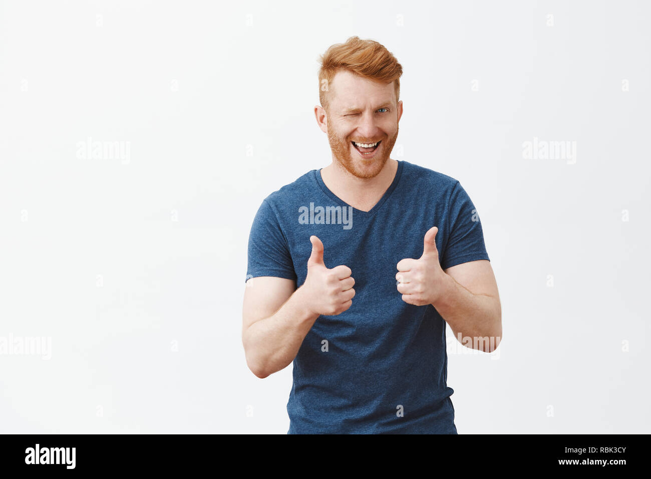 Yeah boy nice choice. Portrait of good-looking male friend with ginger hair showing thumbs up and winking with hint, liking and supporting great decision, cheering over gray background Stock Photo