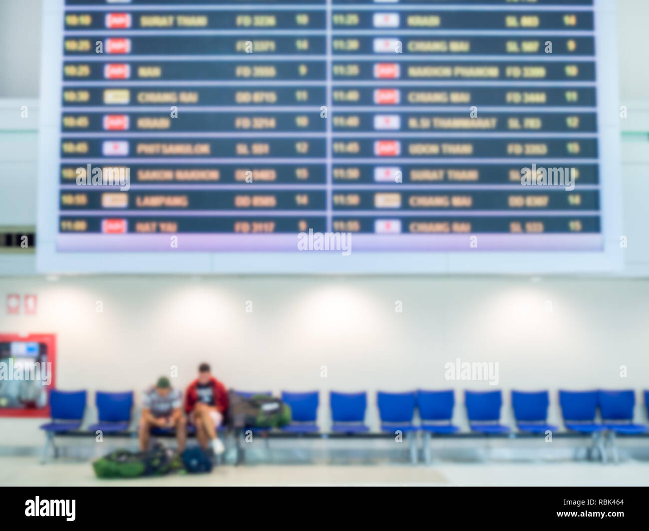 Blurred boarding time monitor screens and passengers sitting on blue chairs in airport hall terminal interior area for arrival passenger. Stock Photo