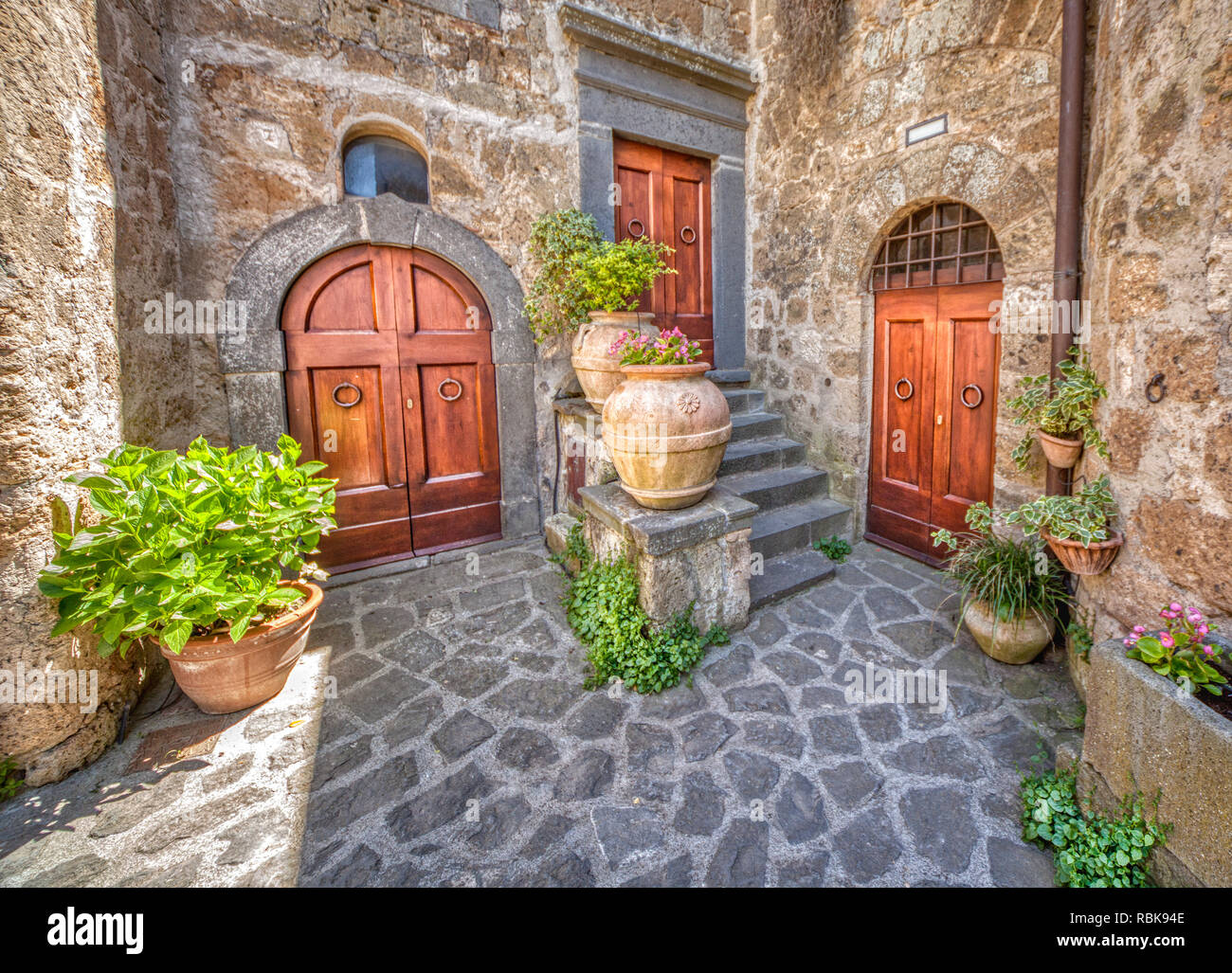 Photograph of a medieval courtyard in Tuscany. Three weathered, worn, brown wood doors of with flowers blooming in clay pots layed on a cobblestone pa Stock Photo