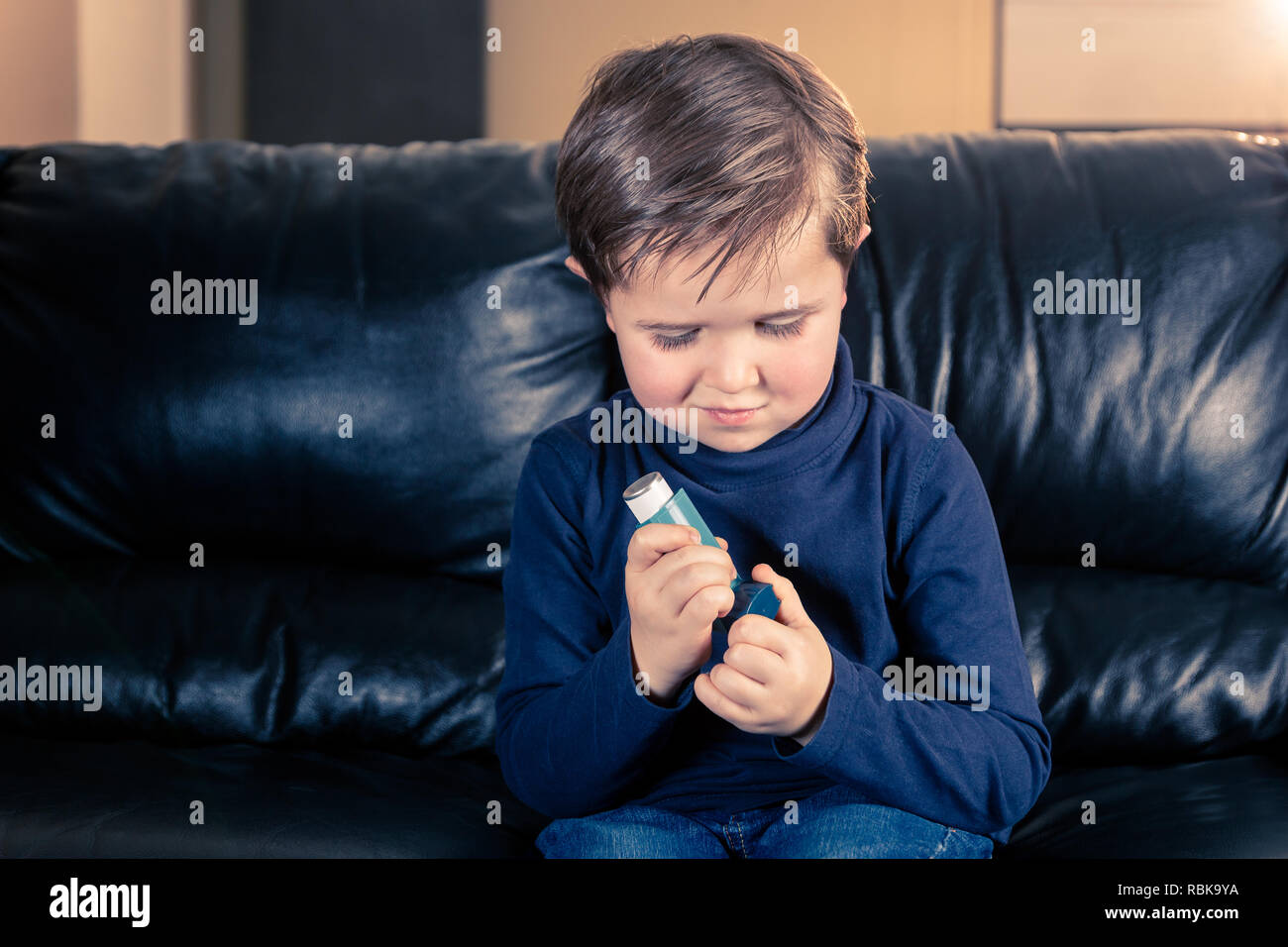 Cute 5 year old boy taking off cap of asthma inhaler, sitting in black leather sofa Stock Photo