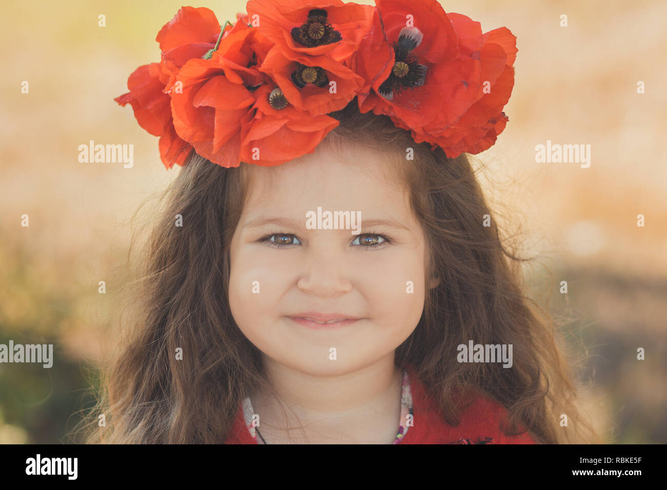 Close portrait of little child girl with big cheeks brown eyes and pout lips wearing stylish red dress with summer shirley poppy on top head. Stock Photo