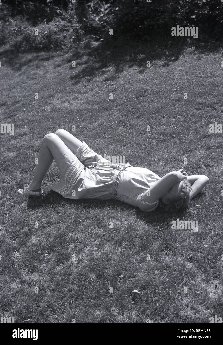 1950s Summertime And A Young Lady Wearing A Light Cotton Dress Lying On The Ground With Her Arm Across Part Of Her Face As If To Say You Can Photo Me But